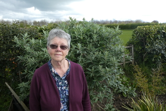 A woman with short grey hair wearing tinted glasses smiles at the camera. There are hedges and green grass in the background and it is cloudy overhead.