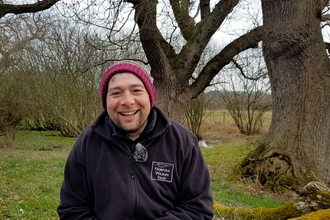 A NWT staff member at Sweet Briar Marshes, smiling at the camera. He is wearing an NWT fleece and a woolly hat.