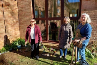 Three people stand in front of a building on a sunny day, holding gardening equipment and smiling at the camera. They are all wearing winter coats and have grey, shoulder length hair