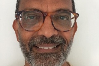 A volunteer smiling at the camera. He has a grey stubbly beard, black hair and round tortoiseshell glasses.