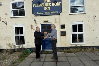 Two men stand in front of the entrance to a pub, which features a sign saying 'The Pleasure Boat Inn'. The men are smiling and looking at each other, while they shake hands with their right hands and exchange a set of keys with their left hands.