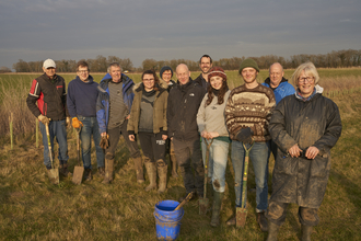 A group of volunteers standing together and smiling at the camera on a sunny winter afternoon. They are wearing wellies and warm clothes, with many holding spades.