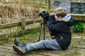 A blond-haired boy wearing a black coat and grey trousers sits on the ground looking through a camera on a tripod