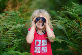 A young girl in a red t-shirt smiles as she looks though a pair of binoculars