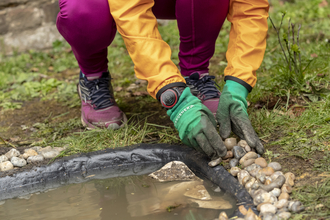 Someone is knelt down, stacking pebbles around the edges of a newly built pond.