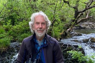 David North posing with his binoculars in front of a small waterfall. He has wavy grey hair and a grey beard.