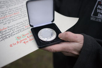 A person's hand holding a silver medal inside a case, with a scroll of paper behind it featuring cursive text.