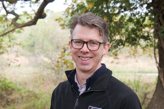 Norfolk Wildlife Trust CEO, Eliot Lyne, smiles at the camera while standing in front of woodland. He is wearing glasses and a black NWT fleece jacket