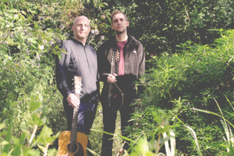 Two men with a guitar and lute stand against a background of green foilage