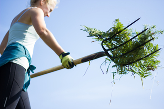 A woman wearing gloves is using a garden fork to gathers cut vegetation for disposal on a sunny, blue skies day. 