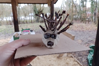 a clay deer with twig antlers held on a piece of cardboard