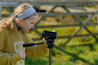 A young girl wearing a yellow sweatshirt looking sideways on at a handheld camera on a tripod