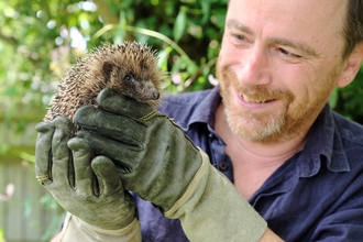 An image of Hugh Warwick wearing green heavy-duty gloves holding a hedgehog