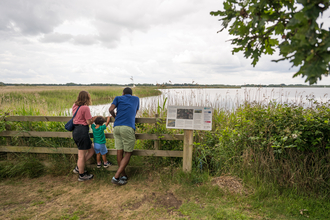 A mum, dad and young boy peering over a fence overlooking the broad at Hickling. 