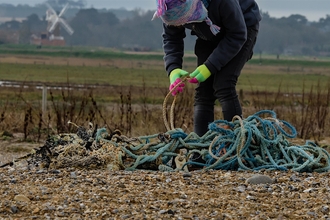 A lady wearing green gloves and a woolly hat bending over and removing rope lying on the beach