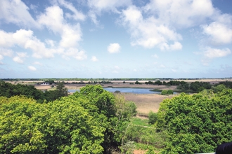 A view over Hickling. Blue skies with fluffy white clouds, lots of trees and shrubs and a pool of water in the distance.