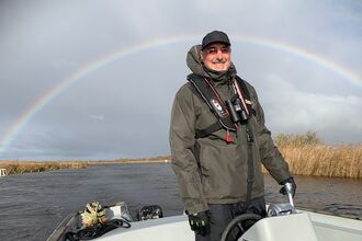A smiling boat operator at Hickling standing in the boat wearing a thick coat and hat and with binoculars around his neck