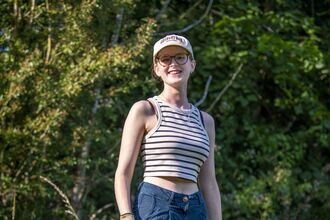 A teenage girl wearing a beige cap, brown glasses and a black and white striped crop top is smiling at the camera.
