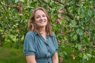 Kate Bradbury standing smiling under a plum tree and wearing a blue dress