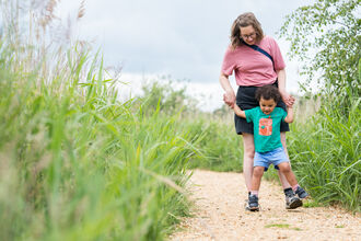 A mum and toddler enjoying a walk at Hickling. She is holding his hands while he stands on her shoes as she walks. 