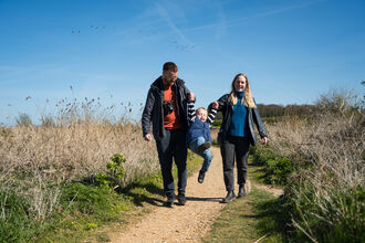 Two parents happily play with their young child as they walk down a path at sunny NWT Cley and Salthouse Marshes nature reserve