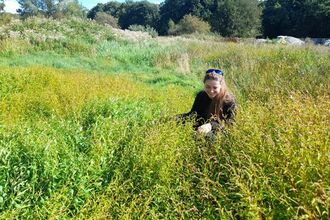 A young woman in a field of tall green plants, grinning on a sunny day. 