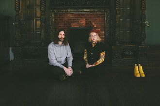Musicians Samantha Whates and MG Boulter are seated in front of a large open fireplace in a dark, historic room