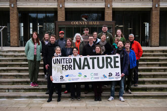 A group of smiling NWT staff on the steps outside Nofolk County Hall holding up a banner that says 'united for nature'.
