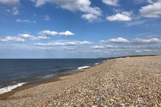A photo of the shingles beach at Cley, with waves lapping on the shore and white clouds in a blue sky
