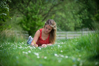 A woman lying on grass covered with daisies, writing. There are trees and bushes visible around her