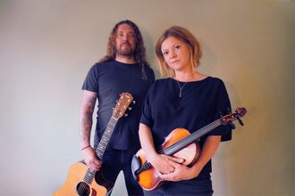 Georgia and Aaren standing together wearing blue t-shirts and holding a guitar and violin