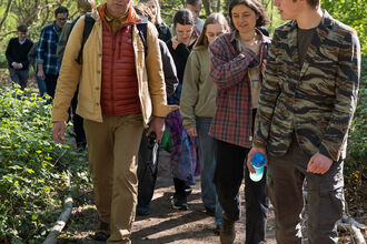 A man wearing a yellow coat and red puffa jacket leads a line of young people in a walk through woodland on a sunny day