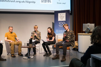Four people sitting in a row on chairs on a stage at an interview panel event. The person on the far right, a teenage boy, is speaking into a microphone.