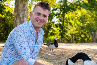 Nick Acheson is wearing a blue and white shirt and blue shorts, smiling at the camera as he crouches down to feed a Canada goose