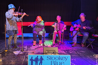 Stookey Blue ceilidh band members in a dark venue playing a fiddle, guitar, flute and accordionn
