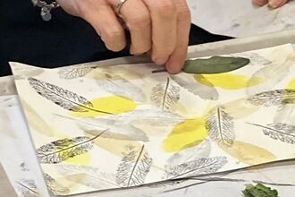a ladies hands creating black, grey and yellow leaf prints on a white piece of paper
