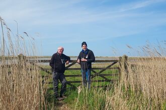 Two men stand in front of a gate in the middle of Cley Marshes. They are both looking at the camera and smiling. They are dressed in black fleeces and combat trousers, with binoculars around their necks.
