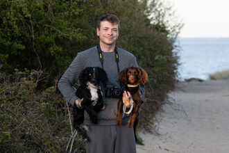 Alfie is standing on a track with his back to the sea. He is holding a spaniel under each arm.