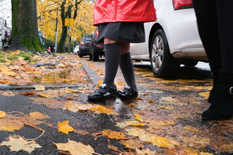 A young girls legs and feet alongside those of an adult, walking on a damp pavement with autumn leaves on the ground