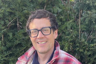 Patrick Barkham is wearing a red and white checked shirt, dark-rimmed glasses and is smiling at the camera