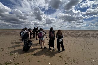 A group of young people, pictured from behind, stand on a beach with a camera on a tripod. It is a sunny day, with thick white clouds dotted around the sky.