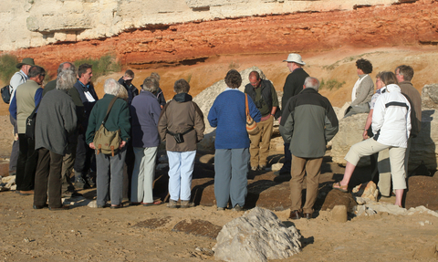 A group of people pictured from behind, gathered at the foot of a cliff. They are standing on sand in front of the orange and yellow cliff wall.