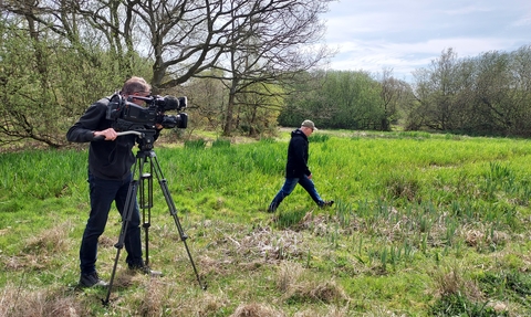 A man stands behind a professional camera on a tripod in a field, while another man walks in front of the camera