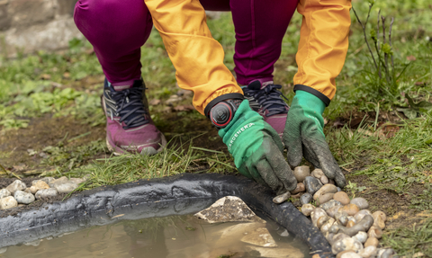 Someone is knelt down, stacking pebbles around the edges of a newly built pond.
