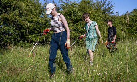 Three young people with gloves, bin bags and pickers, looking for rubbish at Soughbottom Meadows on a sunny day.