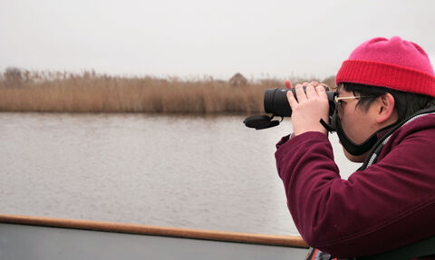 A person dressed in a red coat and hat watches birds through binoculars whilst seated in a small boat on Hickling Broad