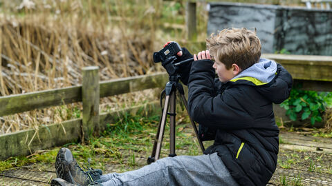 A blond-haired boy wearing a black coat and grey trousers sits on the ground looking through a camera on a tripod