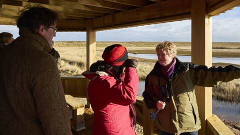 Visitors in coats and hats peering through binoculars at a hide at Cley Marshes on a sunny winter day.