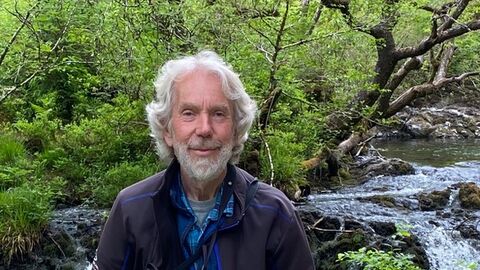 David North posing with his binoculars in front of a small waterfall. He has wavy grey hair and a grey beard.