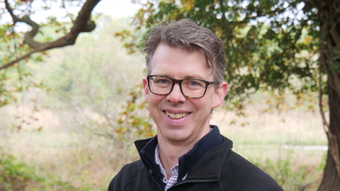Norfolk Wildlife Trust CEO, Eliot Lyne, smiles at the camera while standing in front of woodland. He is wearing glasses and a black NWT fleece jacket